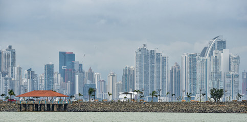 Obraz premium Hot, humid day in Panama city as another rainstorm brews quickly over the city skyline. Tall buildings shimmer in heatwaves rising in humid air. People on Panama Canal jetty park in foreground.