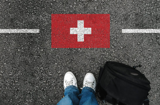  a man with a shoes and backpack is standing on asphalt nex to flag of Switzerland and border
