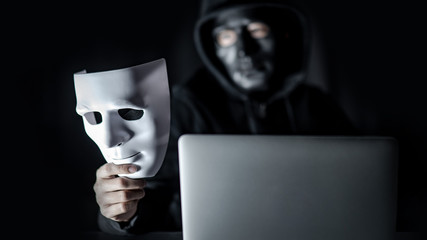 Mystery male hoodie hacker wearing black mask holding white mask sitting with laptop computer on...