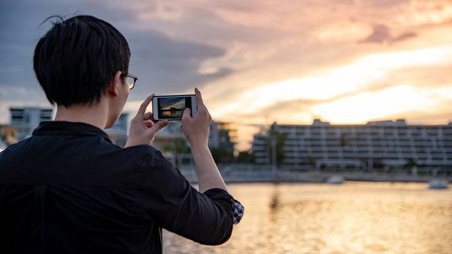 Asian man with glasses using smartphone camera taking photo during the sunset in the city. Urban lifestyle with mobile phone technology concept
