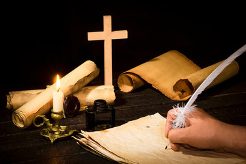 A hand writing with a pen on the background of papyrus scrolls, against the background of a candle and a cross
