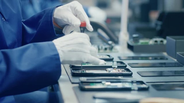 Close Up of a Female Electronics Factory Worker in Blue Work Coat and Protective Glasses Assembling Smartphones with Screwdriver. High Tech Factory Facility with more Employees in the Background. 