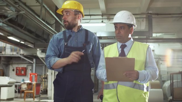 Mid-aged Caucasian technician in overall and hardhat walking with Latin American inspector, telling and showing industrial factory while he writing down on clipboard