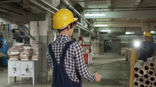 Female technician in overall and hardhat taking cart with paper reels from male coworker and pulling it while working in printing plant