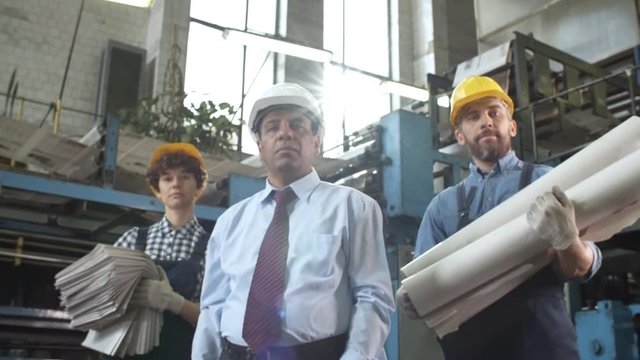 Zoom in of Caucasian male and female technicians in overalls and hardhat holding newspapers and paper reels and Latin American manager standing together in printing plant and posing for camera