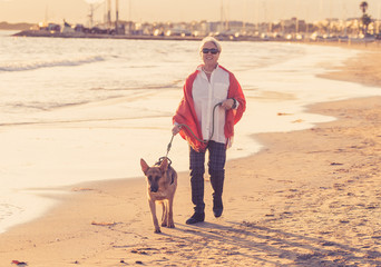 Happy attractive senior woman with her german shepard dog walking on the beach at autumn sunset
