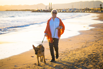 Happy attractive senior woman with her german shepard dog walking on the beach at autumn sunset