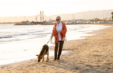 Happy attractive senior woman with her german shepard dog walking on the beach at autumn sunset