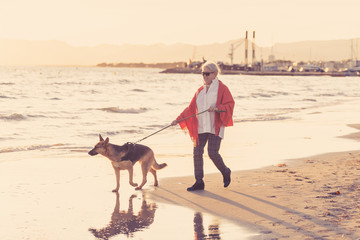 Happy attractive senior woman with her german shepard dog walking on the beach at autumn sunset