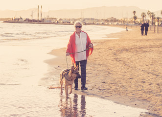 Happy attractive senior woman with her german shepard dog walking on the beach at autumn sunset