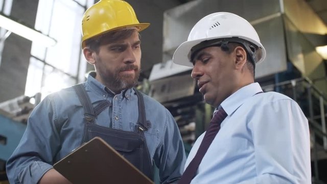 Low angled panning shot of middle aged Latin American manager in hardhat explaining something on clipboard and instructing bearded male worker in industrial plant