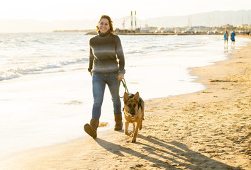 Happy attractive young woman with her german shepard dog walking on the beach at autumn sunset
