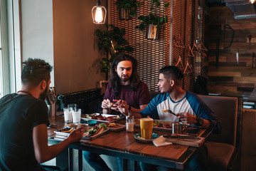 Multiracial friends eat breakfast in cafe. Young men chat while having tasty food and drinks. Guys hangout together