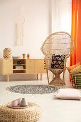 Wicker peacock chair with patterned pillow in bright interior with wooden cabinet and round boho...