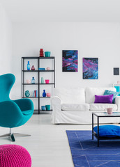 Blue armchair near white settee in living room interior with posters and table on carpet. Real photo