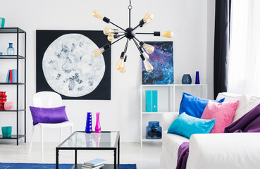 Pink and blue cushions on white couch in living room interior with moon poster behind chair. Real photo