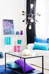 Lamp above table and white settee with blue pillows in living room interior with poster. Real photo