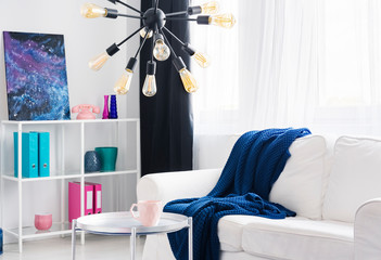 Blue blanket on white couch and silver table under lamp in bright flat interior with poster. Real photo