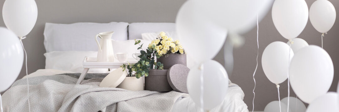 Fresh roses in box and breakfast tray with jug and two cups placed on double bed in real photo with white balloons