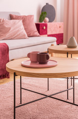 Wooden table with cups on plate on pink carpet in living room interior with grey settee. Real photo