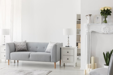 Luxury grey sofa with pillows between two white wooden commodes with stylish lamps, real photo with...