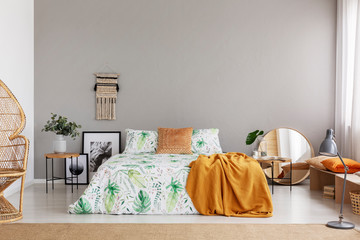 Mustard blanket thrown on king-size bed with leafy sheets in real photo of bright bedroom interior with macrame on the wall and two bedside tables