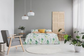 Armchair with houndstooth pattern placed in real photo of bright bedroom interior with two lamps, double bed with leafy sheets and cushions and Monstera Deliciosa plant standing on the floor