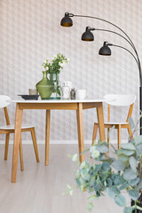 Lamp next to wooden table and white chairs in dining room interior with wallpaper. Real photo