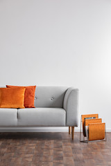 Vertical view of grey comfortable couch with orange pillows and newspaper rack next to it, real photo with copy space on the white wall