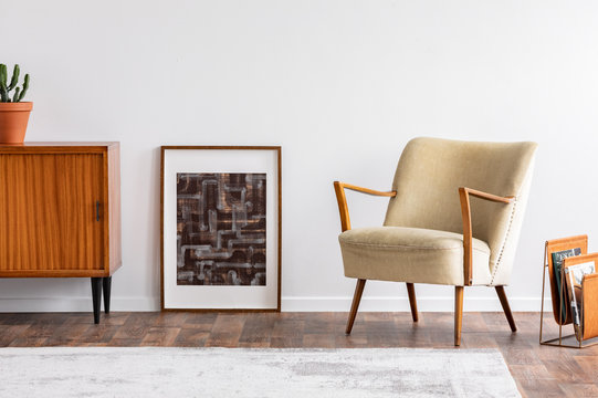 Abstract graphic in wooden frame between retro cabinet with plant and elegant beige armchair, real photo
