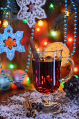 A glass of mulled wine with cinnamon and a slice of orange is on the table against the background of Christmas gingerbread in the shape of snowflakes hanging as Christmas decorations on the branches o