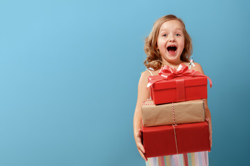 Happy little blonde child girl holding three boxes with gifts on a blue isolated background. The...