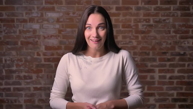Astonished woman watches into the camera and smiles joyfully on a bricken background