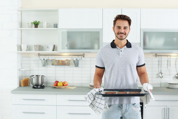 Young man holding oven sheet with cookies in kitchen, space for text