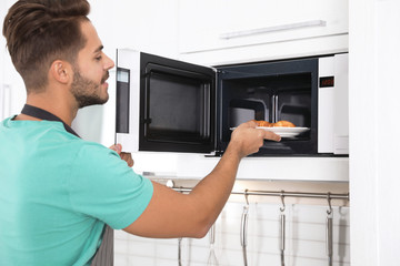 Young man putting plate with croissants into microwave oven at home