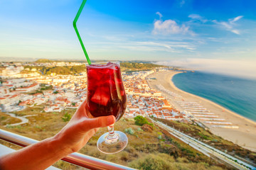 Glass of Sangria in woman hand on Nazare skyline background. Holiday concept and celebration....