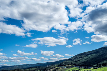 A panoramic and dramatic cloudscape with many clouds on a blue sky over rural mountain hills, with idyllic agricultural farmland, fresh green pasture and forest in sunlight and shades