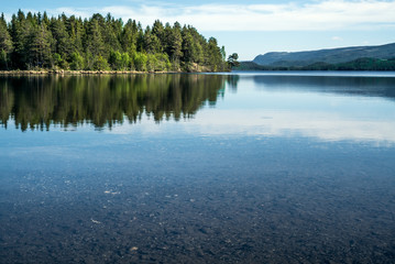 A quiet peaceful summer morning with view over crystal clear calm lake with pebbles in the bottom and green forest reflected on the water surface with mountains in the horizon