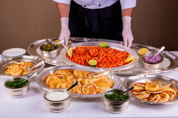 A fish table with pancakes and red fish, sour cream, greens and capers