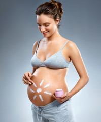 Smiling pregnant woman applying moisturizing cream on her belly on grey background. Pregnancy, maternity, preparation and expectation concept