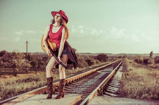 young lady in hat and boots with vintage guitar standing at railways