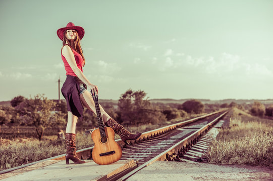 pretty young cowgirl standing at railways with old guitar