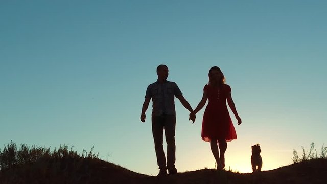 couple silhouette walking. slow motion video. holding hand toward the sun outdoors at sunset dramatic sky background and dog. young happy couple go on nature sunset sun light. man and girl silhouette