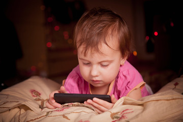 Social Media addiction. Little child girl holding smart phone on the bed before she sleeping at night (psychological problems, media mania, education concept)