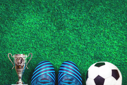 Soccer Ball, Cleats And A Silver Cup Against Green Artificial Turf, Top View With Copy Space