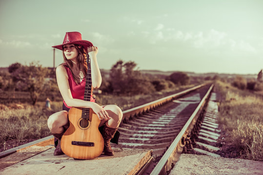 gourgeus brunet in hat with vintage guitar at rural railways sunset