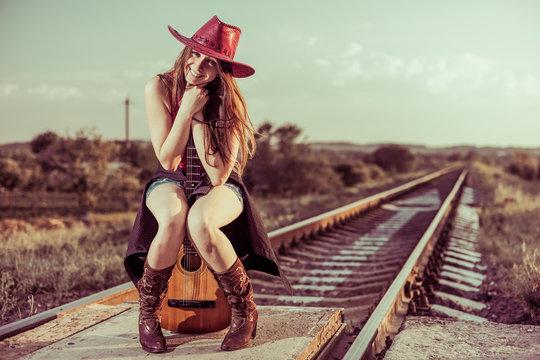 young smiling brunet in hat sitting on guitar at summer railway sunset