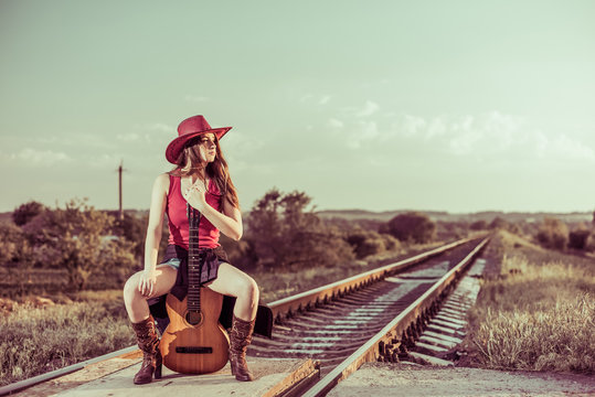 young cowgirl sitting on acoustic guitar at countrysude railways sunset
