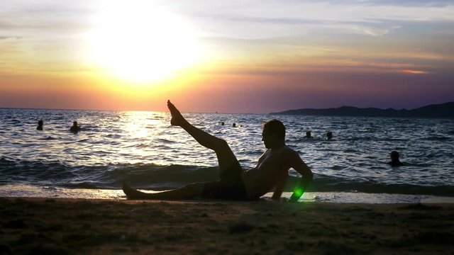 Playful handsome guy in shorts doing fitness, on the seashore against the backdrop of a wonderful sunset