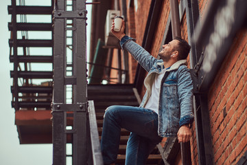 Fototapeta na wymiar A handsome stylish man wearing a denim jacket holding a takeaway coffee on stairs outside.